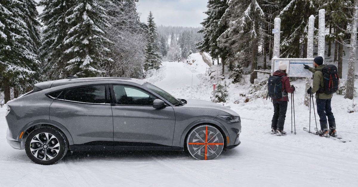 Ein grauer Ford Mustang SUV mit montierten textilen Schneeketten, geparkt auf einer verschneiten Bergstraße, mit Skifahrern in der Nähe unter schneebedeckten Kiefern.