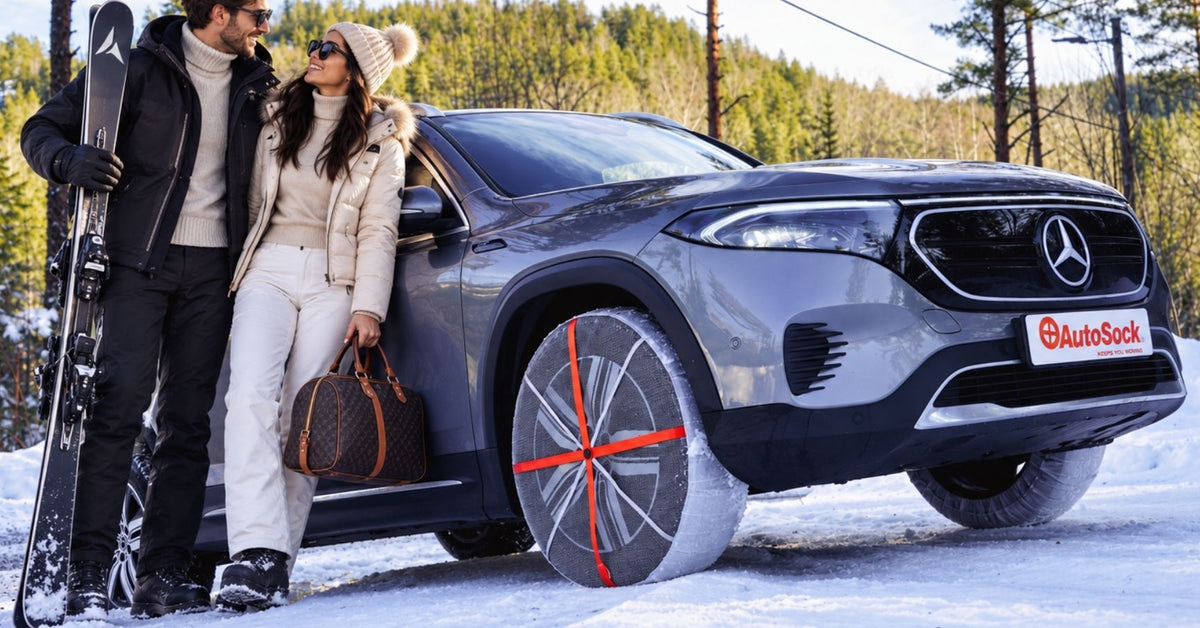 Ein grauer elektrischer Mercedes-Benz SUV mit Schneesocken auf den Winterreifen, geparkt in einer verschneiten Umgebung. Daneben stehen zwei Personen, eine mit Skiern und die andere mit einer braunen Handtasche.